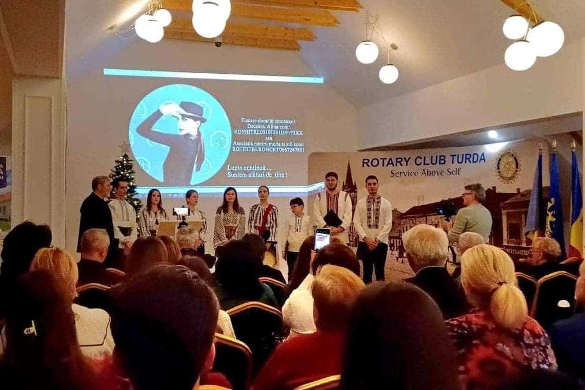 Grupul coral Sf. Ioan Iacob Hozevitul, în costume populare, pe scena de la Salina Gymnasium, în fața bannerului Rotary Club Turda și a unei săli pline